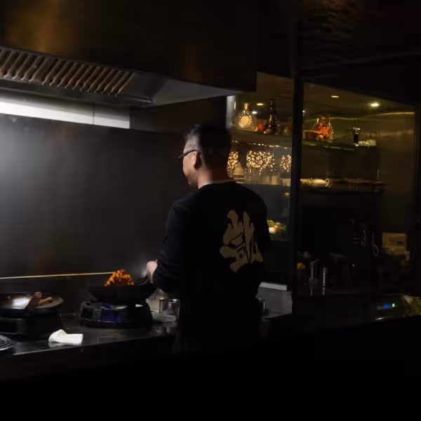 Person cooking food in a dark kitchen with a stove and shelves with glassware in the background.